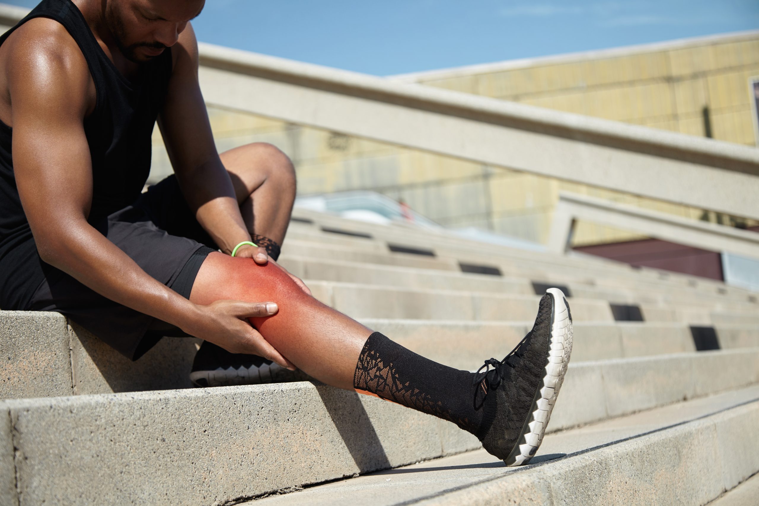 Young sportsman with strong athletic legs holding knee after suffering ligament injury during running workout, sitting on stairs outdoors rubbing red sore area with painful expression on his face ozonoterapia para lesiones deportivas